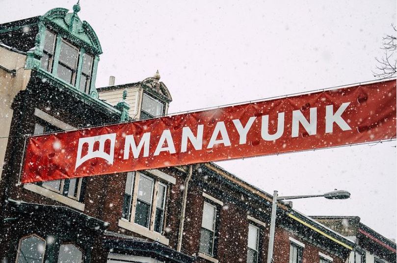 Against a snow filled sky flies a bright red "Manayunk" banner highlighting the beautiful architecture of the deep brick buildings with green and white accents in the small business district. This is a symbol of the community and strength shared among the local Philadelphia business owners and those who support them..